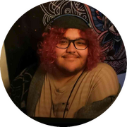 Jazz (a 26 yo, light skinned brown Arab, fat, transman with light stubble) sitting in his gaming chair, wearing his over ear headphones, while smiling at the camera. He has a grey beanie on with shoulder length curly redish pink hair coming out of the side